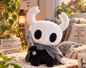May include: A plush toy of the character from Hollow Knight, featuring a white head with two large horns and black oval eyes. The figure wears a dark gray cloak and holds a gray nail-shaped sword. The body is black with rounded limbs.