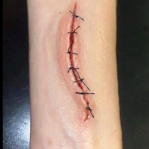 May include: A realistic prosthetic appliance depicting a surgical incision on skin. The prosthetic features a long, vertical wound with deep red coloration in the center, held together by seven distinct black stitches made of thin thread.