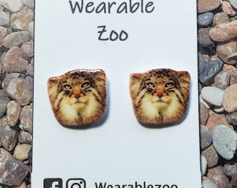May include: A pair of square-shaped earrings featuring a detailed image of a Pallas's cat face. The earrings are set against a white card with the text "Wearable Zoo" and social media icons. The background is a mix of small stones.