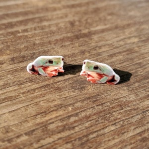 May include: Two frog-shaped earrings. Each earring features a light green body with red and white accents. The frogs have dark eyes and are positioned on a wooden surface.