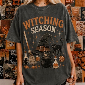May include: A charcoal gray t-shirt featuring the text "WITCHING SEASON" in orange and cream. The graphic includes a black witch hat, a broomstick, a black cat, a bubbling cauldron, two glass potion bottles, a lollipop, and small star accents.