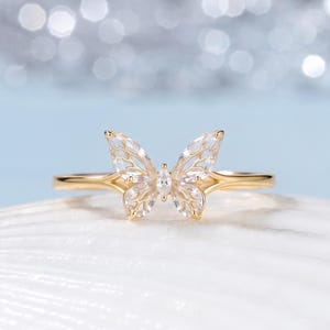 Puede incluir: Un anillo de oro con una pieza central en forma de mariposa adornada con piedras transparentes de talla marquesa. Las alas de la mariposa tienen un diseño calado de estilo filigrana y la banda es de metal dorado pulido y sencillo.