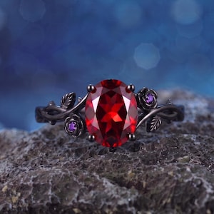 May include: A black metal ring featuring a large oval-cut red gemstone in the center. The band is designed with intricate sculpted rose and leaf motifs, accented by two small round purple stones embedded within the floral metalwork details.