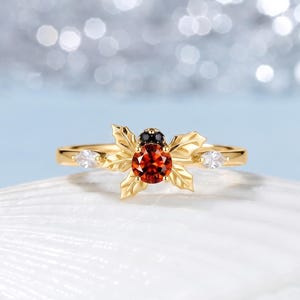 Puede incluir: Un anillo de oro con una piedra preciosa naranja de corte redondo en el centro, rodeada de detalles en forma de hojas doradas. El diseño incluye una pequeña piedra negra en la parte superior y dos piedras claras de corte marquesa a los lados.