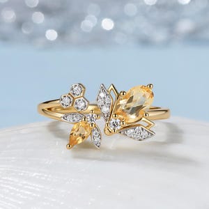 May include: A gold ring featuring a bee motif with two yellow citrine gemstones, one pear-shaped and one oval-shaped, accented by small clear round diamonds set in silver-toned metal, arranged in a honeycomb pattern on a polished gold band.