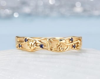 May include: A gold band ring featuring an intricate ocean-themed design with embossed seahorses, jellyfish, and coral motifs. The ring is accented with four small, round, dark blue gemstones set at intervals along the textured, wavy metal surface.