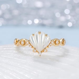 May include: A gold ring featuring a central white shell-shaped inlay, accented with small clear stones along the band and the base of the shell. The band has a twisted metal design that extends from the central shell motif to the sides.