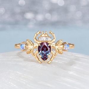 May include: A gold ring featuring a central pear-shaped colour-changing gemstone shaped like a beetle. The beetle design includes a row of small clear stones across the top, leaf-like side accents, and two small round iridescent stones on the band.