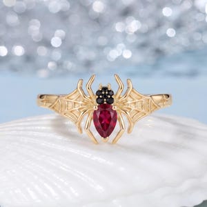 May include: A gold ring featuring a spider design with a pear-shaped red gemstone for the abdomen, small black stones for the head, and spider web patterns extending along the band on both sides of the central spider figure.