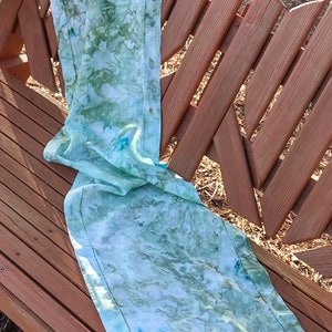 Puede incluir: Un camino de mesa azul claro y verde con fondo blanco y un patrón sutil. El camino de mesa está doblado y colocado en un banco de madera.