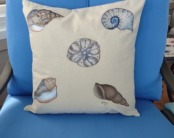 May include: Square, cream-coloured cushion featuring hand-painted seashells in shades of blue, brown, and grey. The cushion has five different seashell designs. The cushion is on a blue chair.