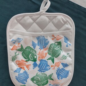 May include: White quilted oven mitt with a loop for hanging. The mitt features a pattern of green, blue, and orange fish. The mitt has a white border and a small pocket.