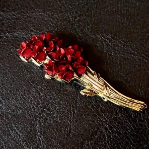 May include: A gold-tone metal brooch shaped like a bouquet of flowers with deep red petals. The design features a cluster of layered red blossoms at the top, connected to a slender, textured gold stem with a small leaf detail extending from the side.