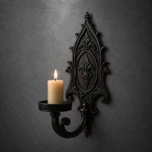 May include: A black wall-mounted candle holder with an ornate, pointed backplate featuring intricate scrollwork and a single red gemstone accent. A cylindrical cream-colored pillar candle sits in the circular holder, with a lit flame at the top.