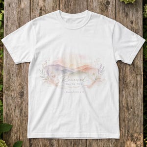 Könnte beinhalten: Ein weißes T-Shirt mit kurzen Ärmeln und einer Aquarellgrafik von Bergen, einer Sonne und floralen Elementen. Der Text "Renewed Day by Day" sowie "2 Corinthians 4:16" sind darunter in einer Schreib- und Serifenschrift gedruckt.