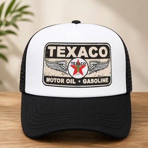 Può includere: Un cappellino trucker bianco e nero con una grafica rettangolare sul pannello frontale. La grafica mostra la parola TEXACO in lettere in grassetto sopra un logo a stella rossa con ali, e il testo MOTOR OIL e GASOLINE sul bordo inferiore.