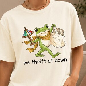 Puede incluir: Una camiseta de manga corta color crema con el gráfico de una rana verde con bufanda amarilla, que lleva una lámpara de vidrieras y una bolsa de la compra blanca. Debajo de la ilustración, el texto dice we thrift at dawn en letras negras.