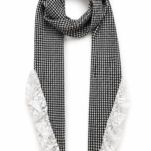 May include: A long scarf featuring a black and white houndstooth pattern throughout the main body. The two ends of the scarf are finished with a decorative white lace trim that runs along the outer edges, creating a ruffled effect on both sides.
