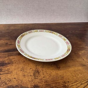 May include: A round white ceramic plate featuring a decorative border pattern. The rim is adorned with a repeating design of small pink floral clusters and yellow scrollwork accents, separated by tiny dark geometric diamond shapes.