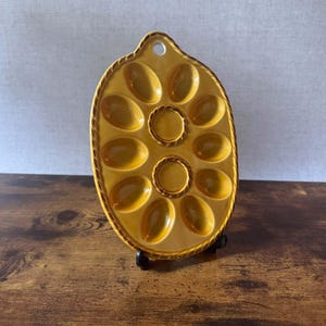 May include: An oval-shaped mustard yellow ceramic egg serving tray featuring ten recessed egg-shaped indentations arranged around two central circular wells. The tray has a rope-textured border and a single hole at the top for hanging.