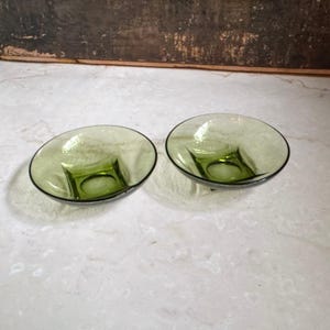 Può includere: Due piccole ciotole rotonde realizzate in vetro verde oliva traslucido. Ogni ciotola presenta una base quadrata che si trasforma in un bordo circolare, creando un profilo liscio e curvo. Il vetro ha un aspetto sottilmente strutturato.