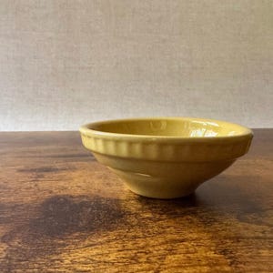 May include: A mustard yellow ceramic bowl with a smooth, glossy finish and a distinct ribbed texture along the outer rim. The bowl features a wide, open top that tapers down to a smaller, rounded base, showcasing a simple, functional design.