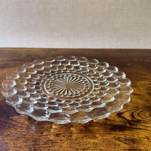 May include: A clear glass plate featuring a textured surface with a repeating pattern of raised circular dots arranged in concentric rings around a central sunburst design with radiating lines, creating a decorative and reflective appearance.