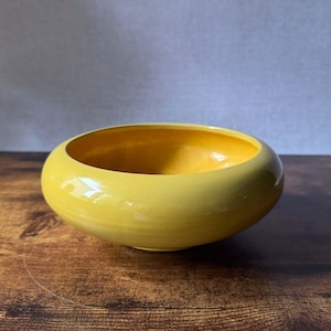May include: A bright yellow ceramic bowl with a smooth, glossy finish and a wide, rounded rim. The bowl features a deep, circular basin and a low-profile design, showcasing a vibrant, solid color throughout its entire exterior and interior surface.