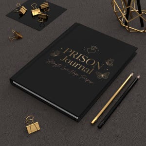 Könnte beinhalten: Ein schwarzes Notizbuch mit goldenem Text PRISON JOURNAL und Strength Love Hope Purpose, verziert mit goldenen Schmetterlings- und Herzsymbolen. Der Einband zeigt eine Krone. Zwei Stifte und mehrere goldene Büroklammern liegen daneben.