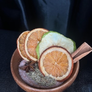 Puede incluir: Un pequeño cuenco de madera lleno de sal rosa, romero seco y un anís estrellado. El cuenco está cubierto con tres rodajas de naranja seca, una rodaja de manzana verde seca y una rama de canela, creando un arreglo texturizado y aromático.