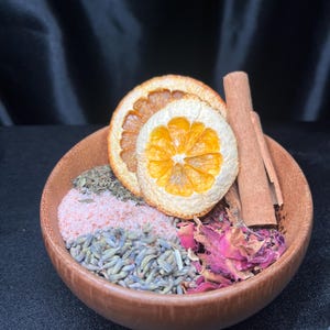 May include: A round wooden bowl filled with a mixture of dried ingredients including two orange slices, two cinnamon sticks, pink salt, dried lavender buds, dried rose petals, and green herbs arranged in distinct sections within the bowl.