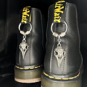 Könnte beinhalten: Ein Paar schwarze Lederstiefel mit gelben Nähten entlang der Sohle und einer gelben AirWair-Fersenschlaufe. Jeder Stiefel ist mit einem silberfarbenen Metallring an der Schlaufe verziert, der einen Anhänger in Form eines Vogelschädels trägt.