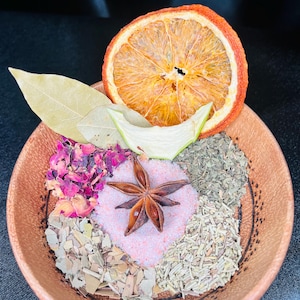 Puede incluir: Un cuenco redondo y texturizado de color marrón lleno de diversos ingredientes botánicos secos, incluyendo una rodaja de naranja seca grande, hojas de laurel, sal rosa, un anís estrellado, pétalos de rosa secos y varias hierbas verdes.