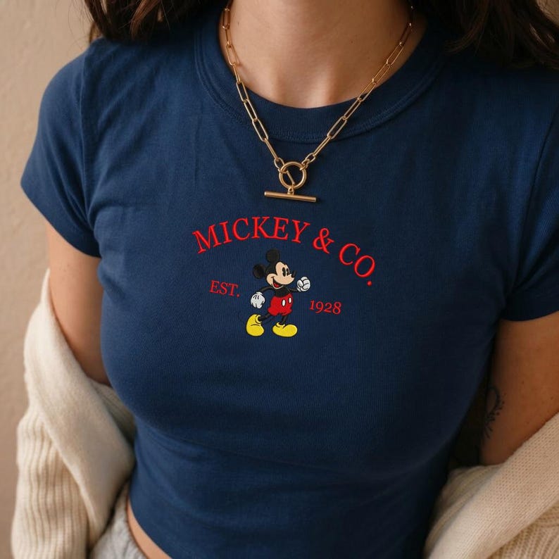 May include: A navy blue short-sleeved t-shirt featuring a red embroidered graphic that reads MICKEY & CO. EST. 1928 with a central illustration of Mickey Mouse. A gold-toned chain necklace with a toggle clasp is worn around the neck.