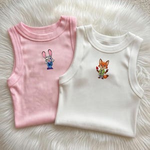 May include: Two ribbed tank tops, one light pink and one white, each featuring a small embroidered character on the chest. The pink tank displays a grey rabbit character, while the white tank features an orange fox character holding a red popsicle.