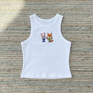 May include: A white sleeveless ribbed tank top featuring a small, centered graphic print of a cartoon rabbit and a cartoon fox standing side by side. The rabbit has grey fur and a blue uniform, while the fox has orange fur, a green shirt, and a tie.