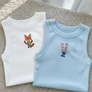 May include: A white ribbed tank top featuring an embroidered orange fox character holding a red popsicle and a light blue ribbed tank top featuring an embroidered grey rabbit character with large ears and a blue uniform, both with crew necklines.