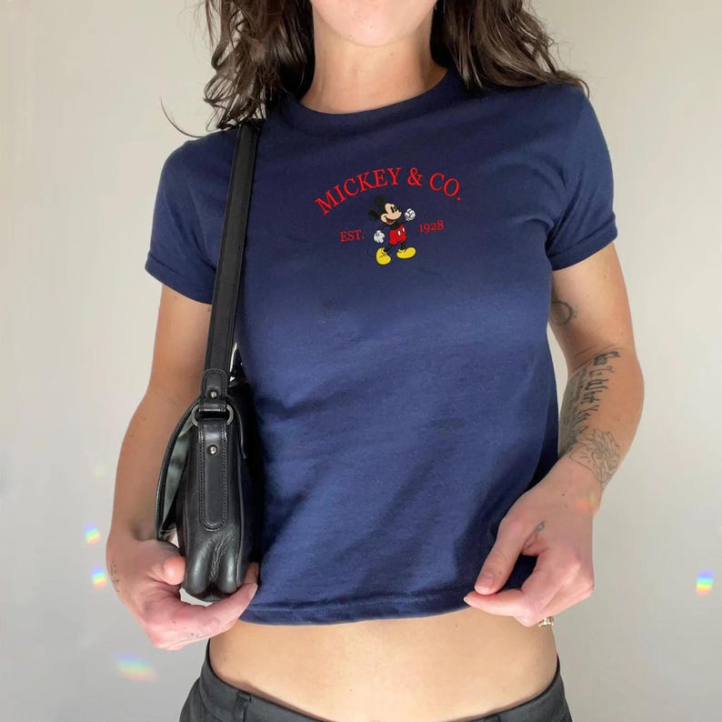 May include: A navy blue short-sleeved t-shirt featuring a graphic of Mickey Mouse and the red text MICKEY & CO. EST. 1928. A black shoulder bag with a long strap is positioned at the side of the shirt, which has a crew neckline and a fitted cut.