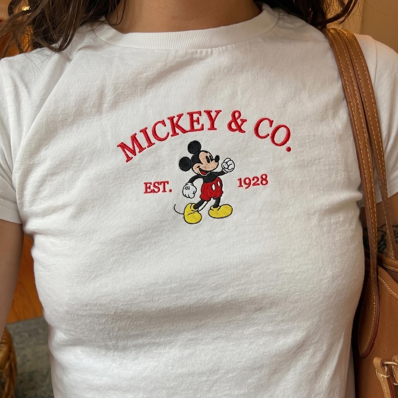 May include: A white short-sleeved t-shirt featuring an embroidered graphic of Mickey Mouse in the center. The text MICKEY & CO. is arched above the character in red, with EST. 1928 printed in red on either side of the figure in a classic font.