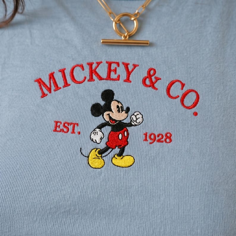 May include: A light blue t-shirt featuring an embroidered graphic of Mickey Mouse in red shorts and yellow shoes. The text MICKEY & CO. is arched above the character, with EST. 1928 printed below. A gold-toned metal toggle necklace rests above.