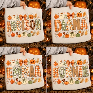 Può includere: Una felpa di colore chiaro con un nome personalizzato in grandi lettere maiuscole riempite di motivi autunnali come tartan, fiori e pois. Il design include zucche, foglie d'acero, un fantasma e un fiocco decorativo.