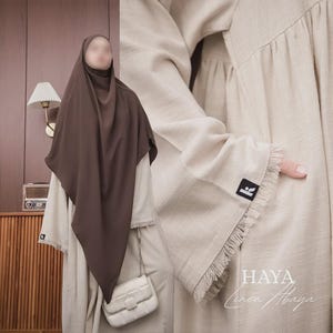 Peut inclure: Une abaya en lin beige avec des manches longues aux bords effilochés et une petite étiquette noire de marque. Un hijab marron foncé couvre la tête et les épaules, accompagné d'un sac à bandoulière matelassé blanc. Le texte HAYA Linen Abaya est visible.