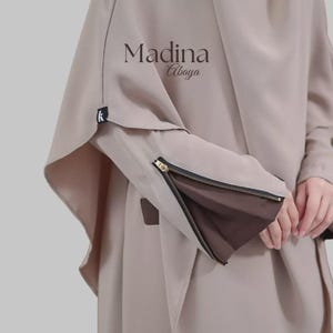 May include: A light beige abaya featuring the text Madina Abaya printed in dark brown. The sleeve has a functional gold-toned zip that opens to reveal a dark brown fabric lining, and a small black fabric tag with a white letter k is attached.