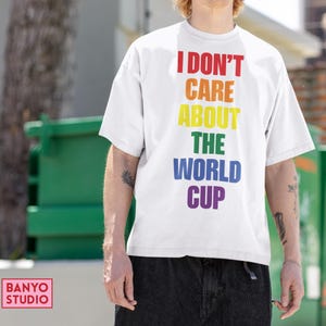 Op de afbeelding: Een wit T-shirt met korte mouwen en de tekst I DON'T CARE ABOUT THE WORLD CUP verticaal gedrukt in een regenboogkleurenschema, waarbij elk woord een andere kleur heeft, waaronder rood, oranje, geel, groen, blauw en paars.