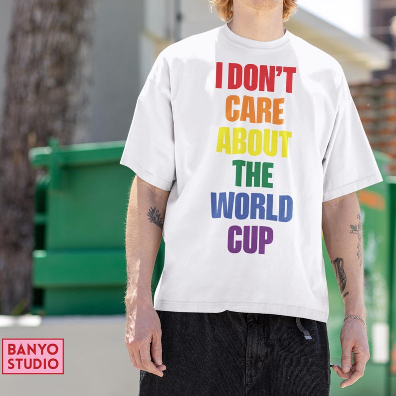 Op de afbeelding: Een wit T-shirt met korte mouwen en de tekst I DON'T CARE ABOUT THE WORLD CUP verticaal gedrukt in een regenboogkleurenschema, waarbij elk woord een andere kleur heeft, waaronder rood, oranje, geel, groen, blauw en paars.