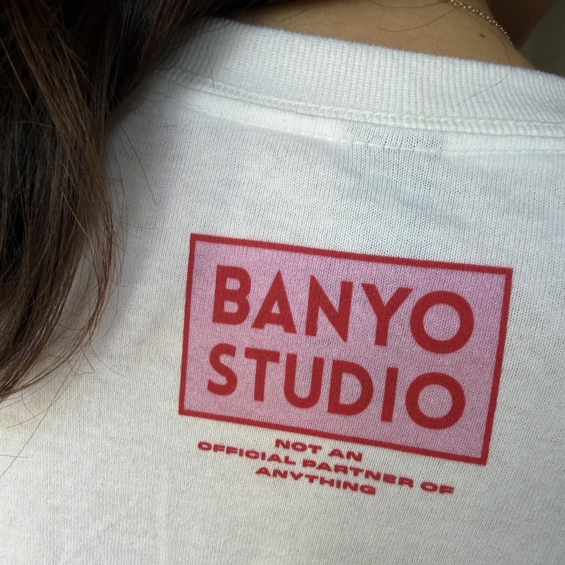 Op de afbeelding: Een wit t-shirt met een rechthoekige roze afbeelding met de rode tekst BANYO STUDIO. Onder de rechthoek staat in kleinere rode letters NOT AN OFFICIAL PARTNER OF ANYTHING. Het ontwerp bevindt zich op de bovenrug van het kledingstuk.