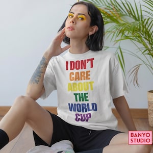 Op de afbeelding: Een wit T-shirt met korte mouwen en de tekst I DON'T CARE ABOUT THE WORLD CUP in een verticaal regenboogverloop van rode, oranje, gele, groene, blauwe en paarse hoofdletters, gecentreerd op de voorzijde van het kledingstuk.