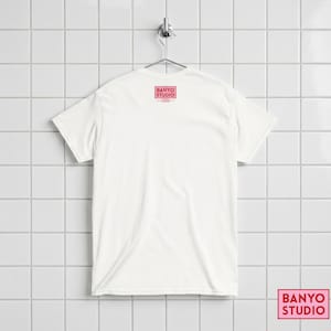 Op de afbeelding: Een effen wit t-shirt met korte mouwen dat aan een metalen hanger hangt. De achterkant van het shirt heeft een klein rood rechthoekig logo bij de halslijn met de tekst BANYO STUDIO in een vet schreefloos lettertype, met kleinere tekst eronder.