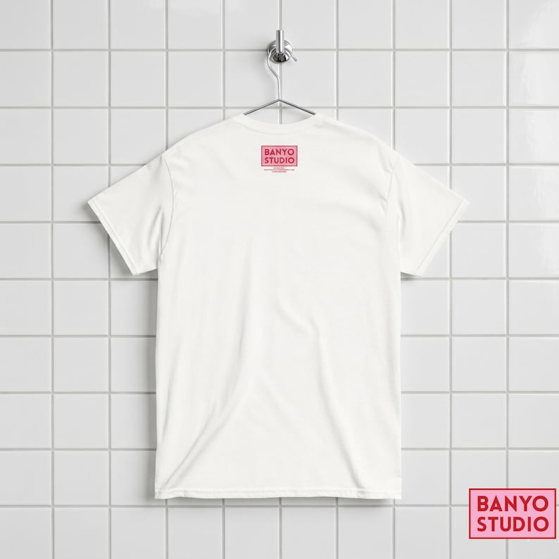 Op de afbeelding: Een effen wit t-shirt met korte mouwen dat aan een metalen hanger hangt. De achterkant van het shirt heeft een klein rood rechthoekig logo bij de halslijn met de tekst BANYO STUDIO in een vet schreefloos lettertype, met kleinere tekst eronder.