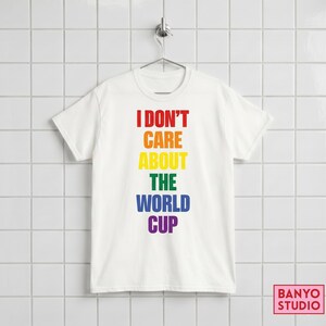 Op de afbeelding: Een wit t-shirt met korte mouwen en de tekst I DON'T CARE ABOUT THE WORLD CUP in een verticaal regenboogverloop van rode, oranje, gele, groene, blauwe en paarse hoofdletters, gecentreerd op de voorzijde van het kledingstuk.